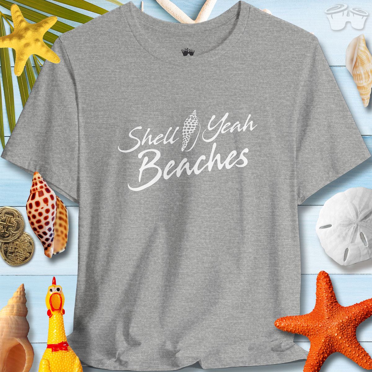 Gray short sleeve tee with 'Shell Yeah Beaches' text, surrounded by seashells and beach decor, promoting summer vibes.