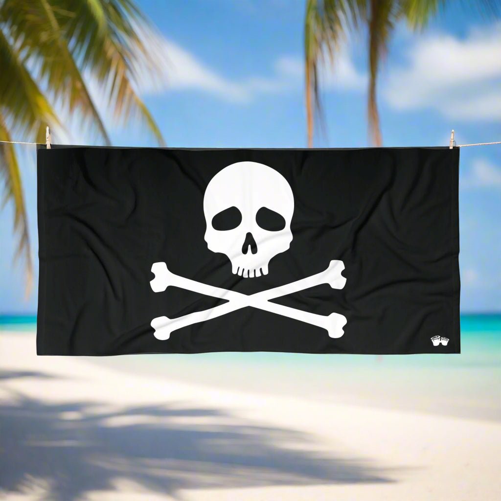 Jolly Roger Pirate Flag Beach Towel with skull and crossbones design hanging on a line in a tropical beach paradise