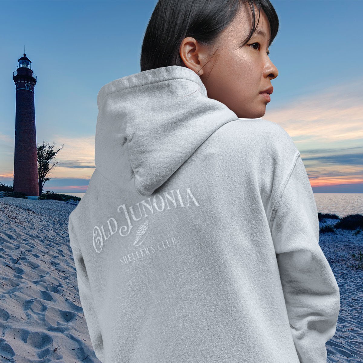 Person wearing a gray hoodie with 'Old Junonia' text on a beach with a lighthouse in the background.