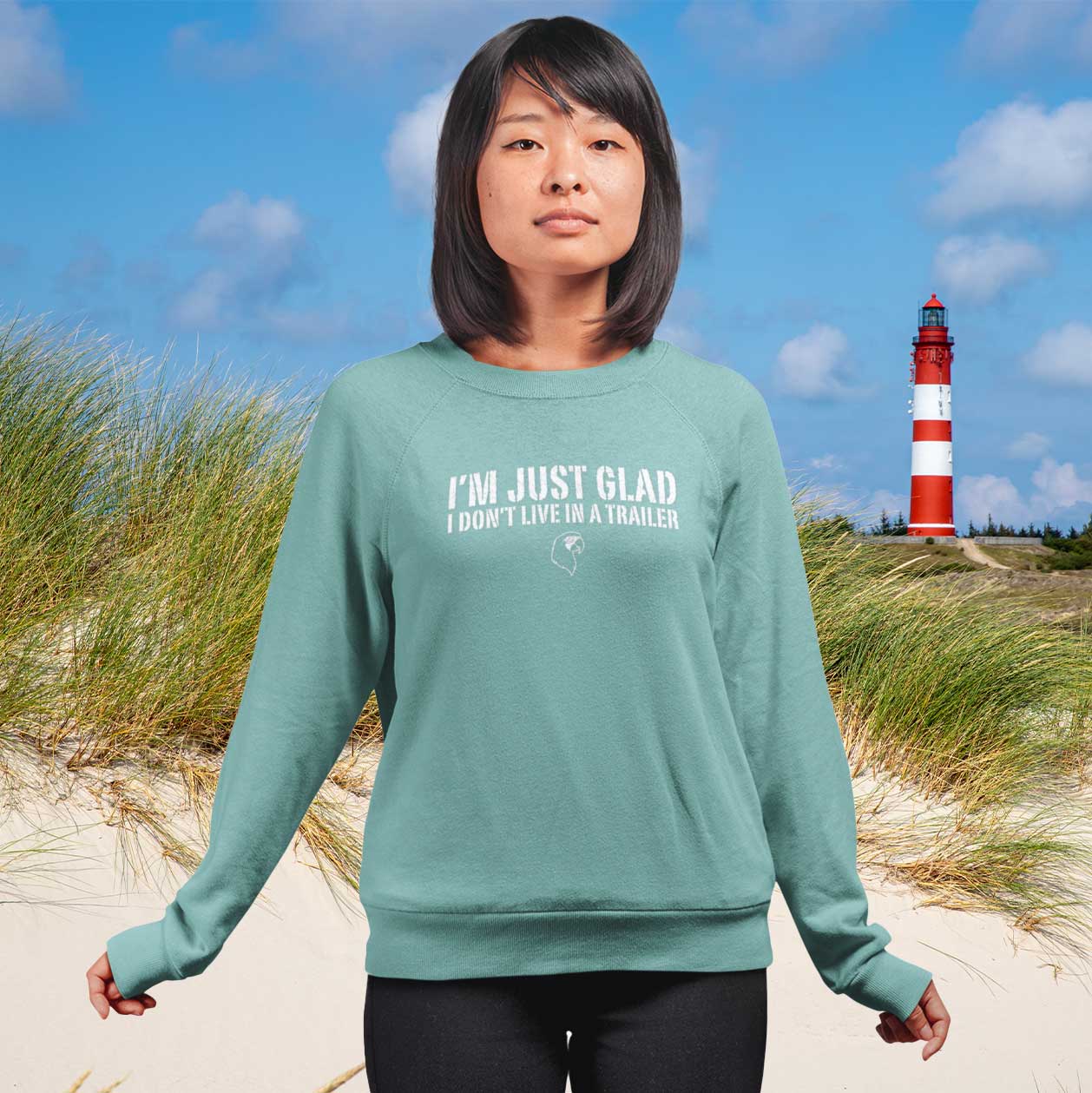 Person wearing a teal sweatshirt with text on a beach with a lighthouse in the background