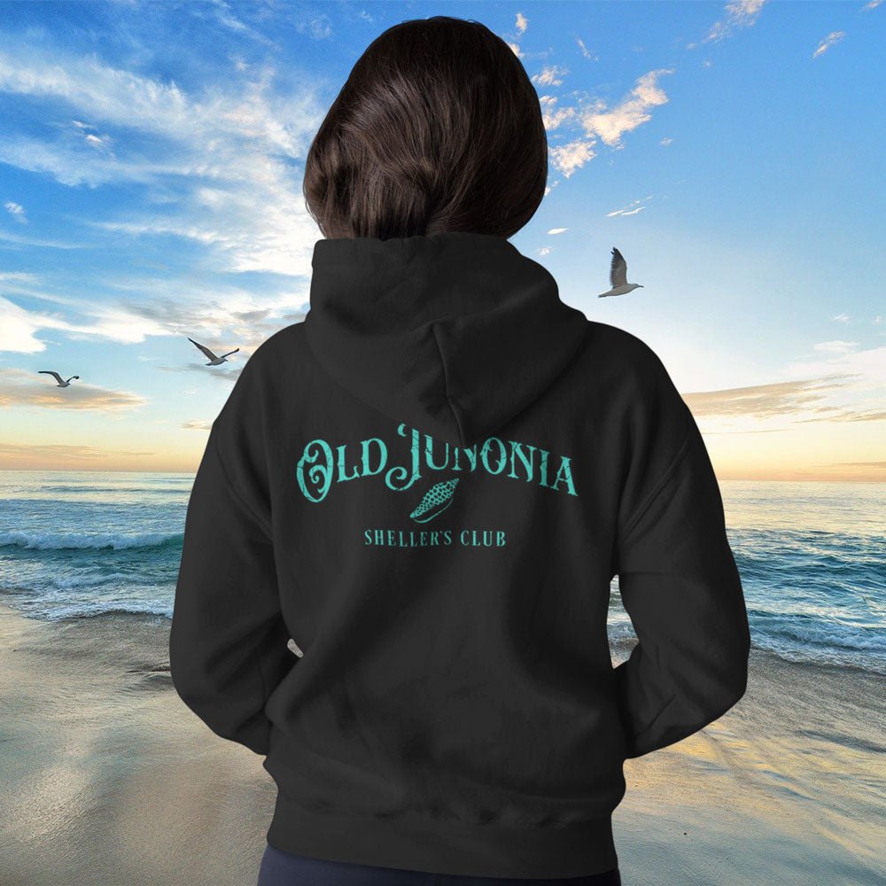 Person wearing a black hoodie with 'Old Jutlandia Sheller's Club' text on the beach at sunset.