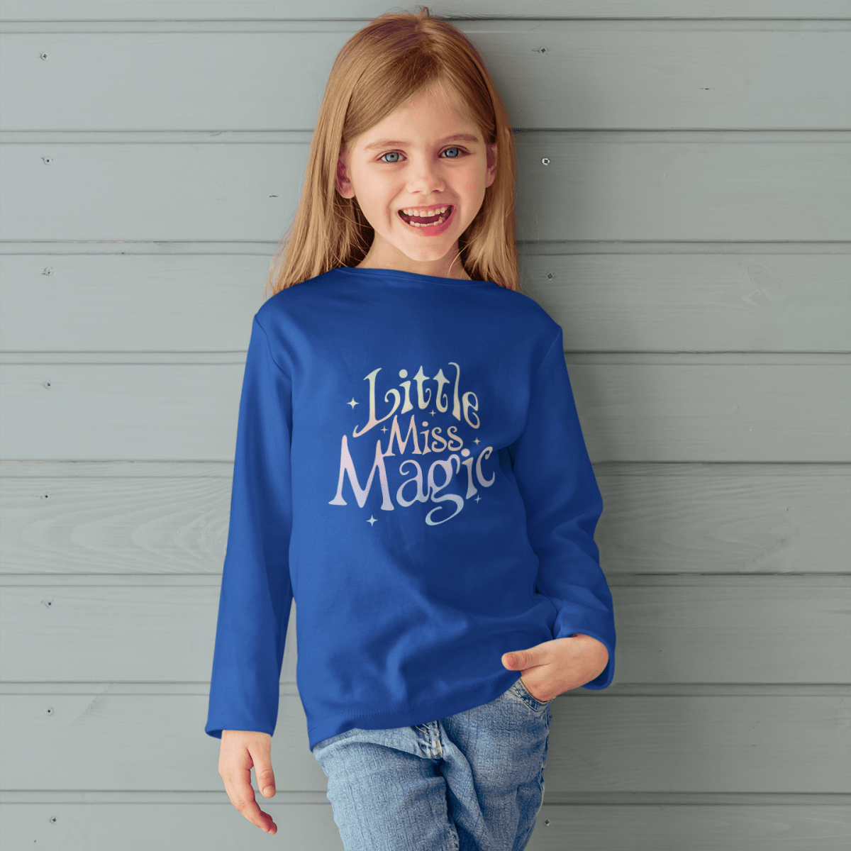 Child wearing a blue sweatshirt with 'Little Miss Magic' text against a gray wooden wall.