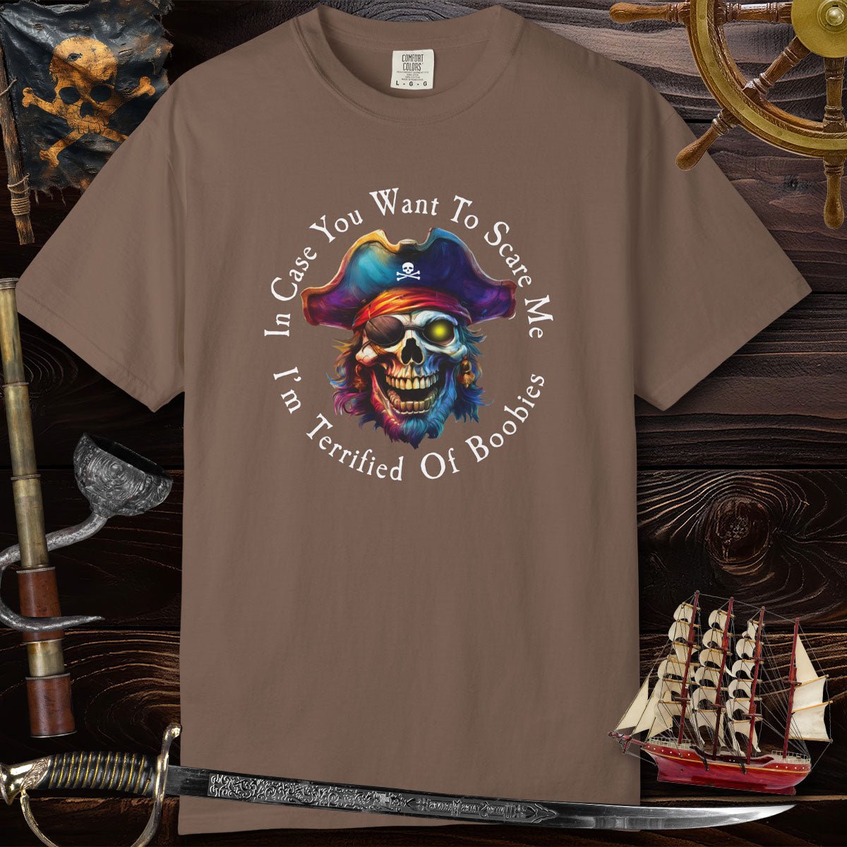 Brown t-shirt with a colorful pirate skull design and text, placed on a wooden background with ship and sword.