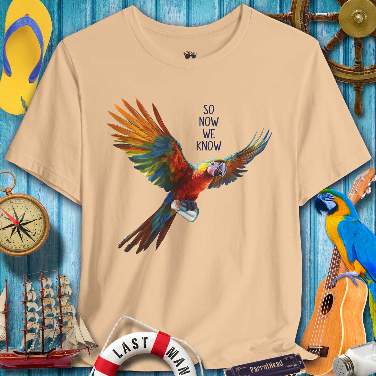 Beige t-shirt with a colorful parrot graphic and text on a blue wooden background with nautical items.