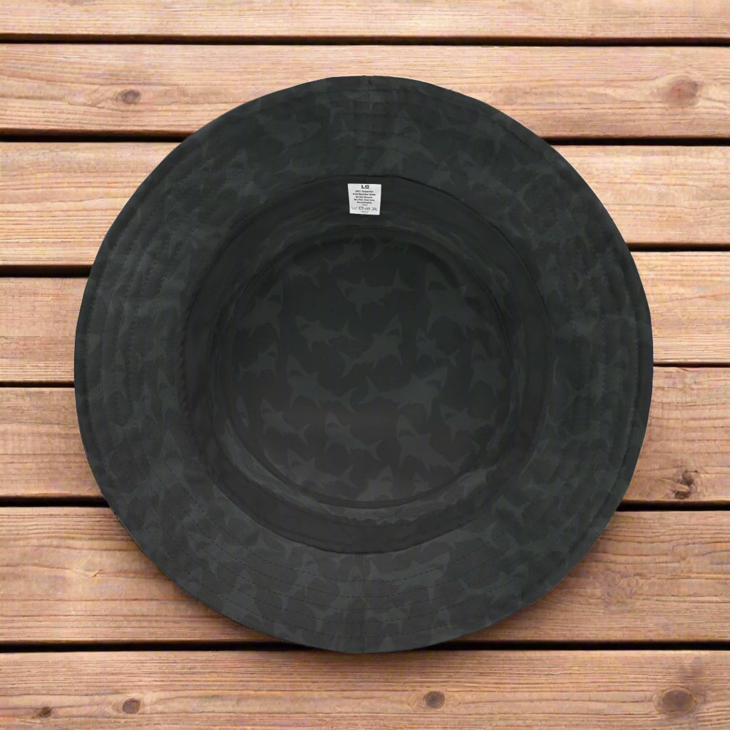 Underside of a black bucket hat with a subtle shark pattern lying on a rustic wood deck.