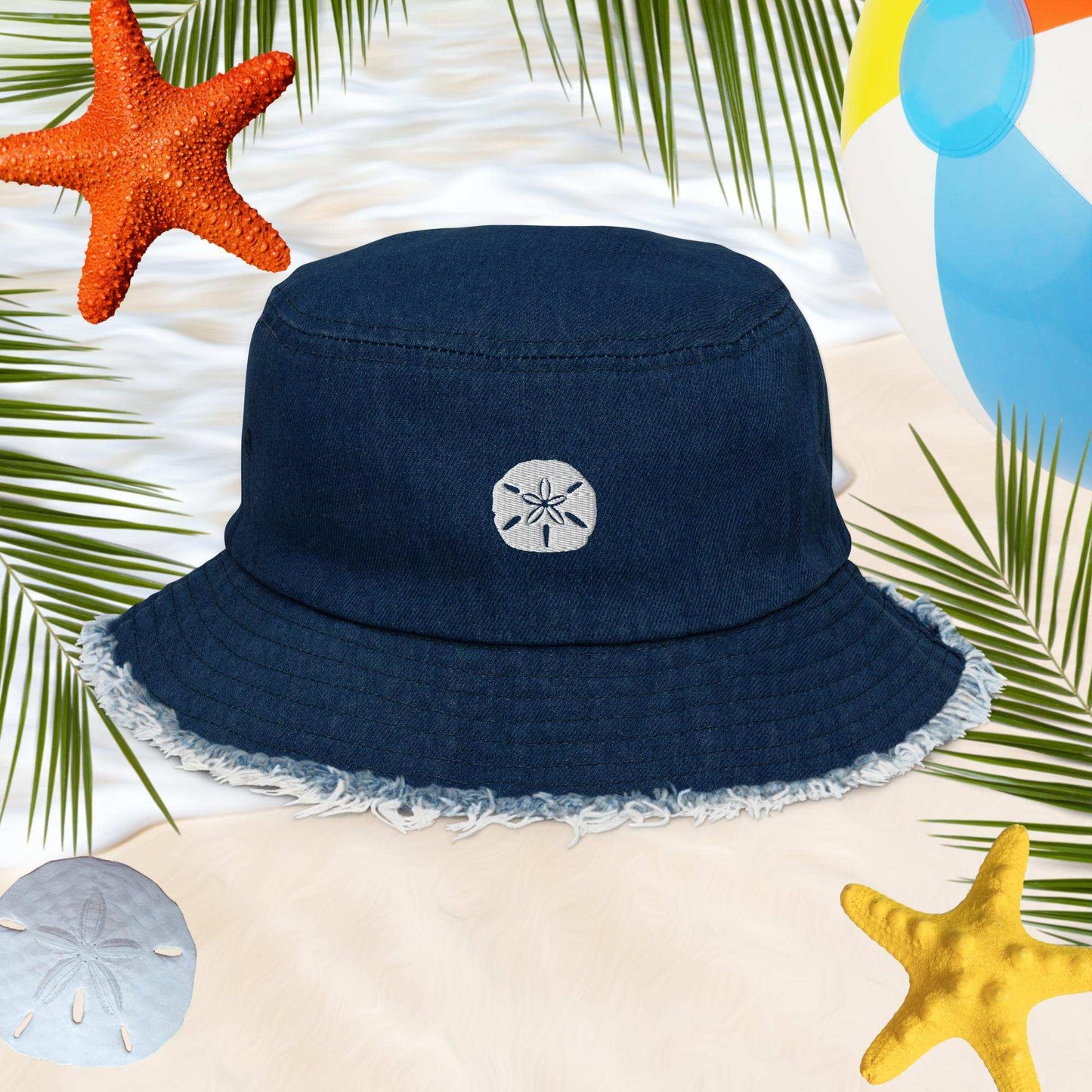 Dark Blue Denim Bucket Hat with Sand Dollar Embroidery and frayed edges around the brim. In the background is a beach with a sand dollar some starfish and a beachball.
