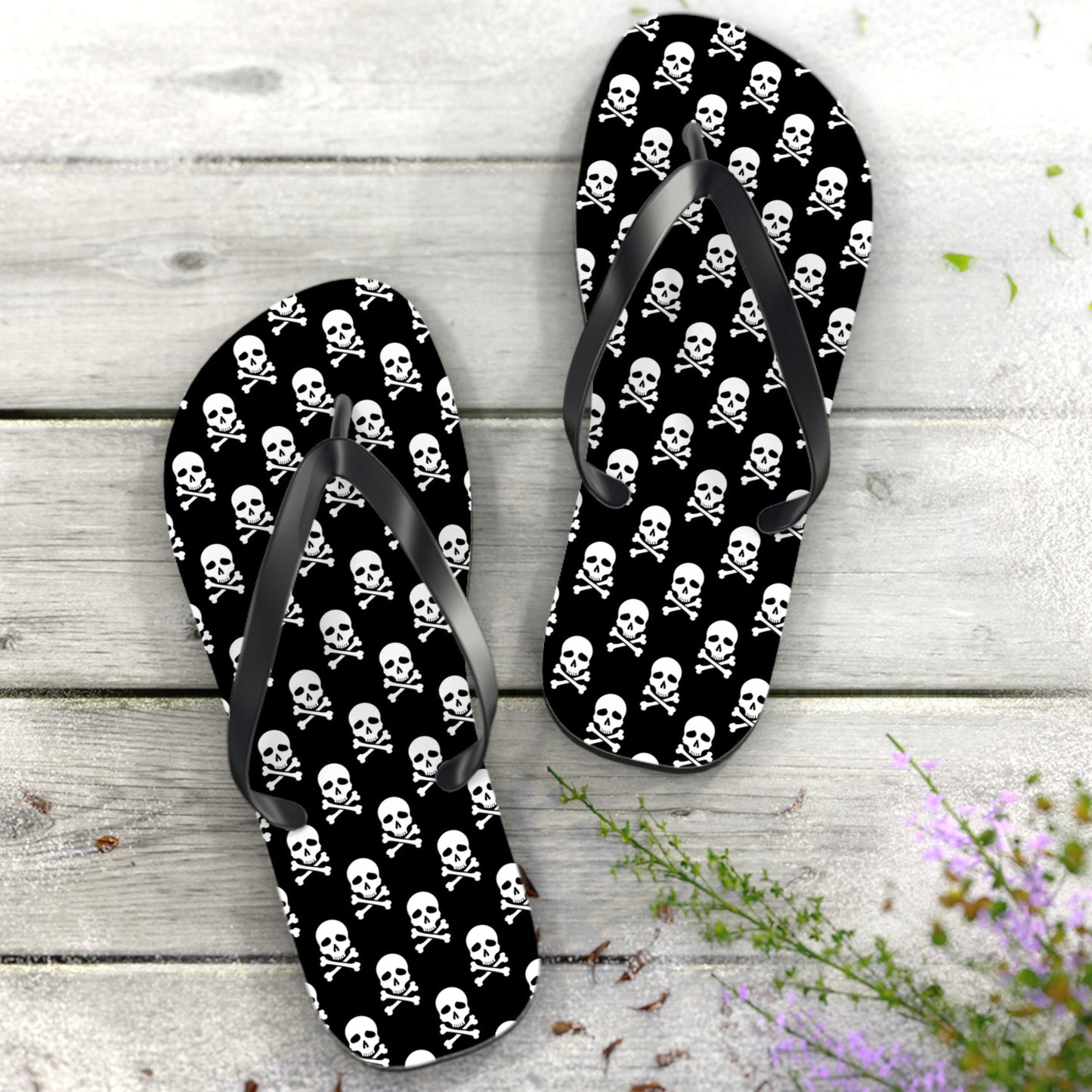 Flip-flops with skull and crossbones pattern on a wooden surface with flowers.