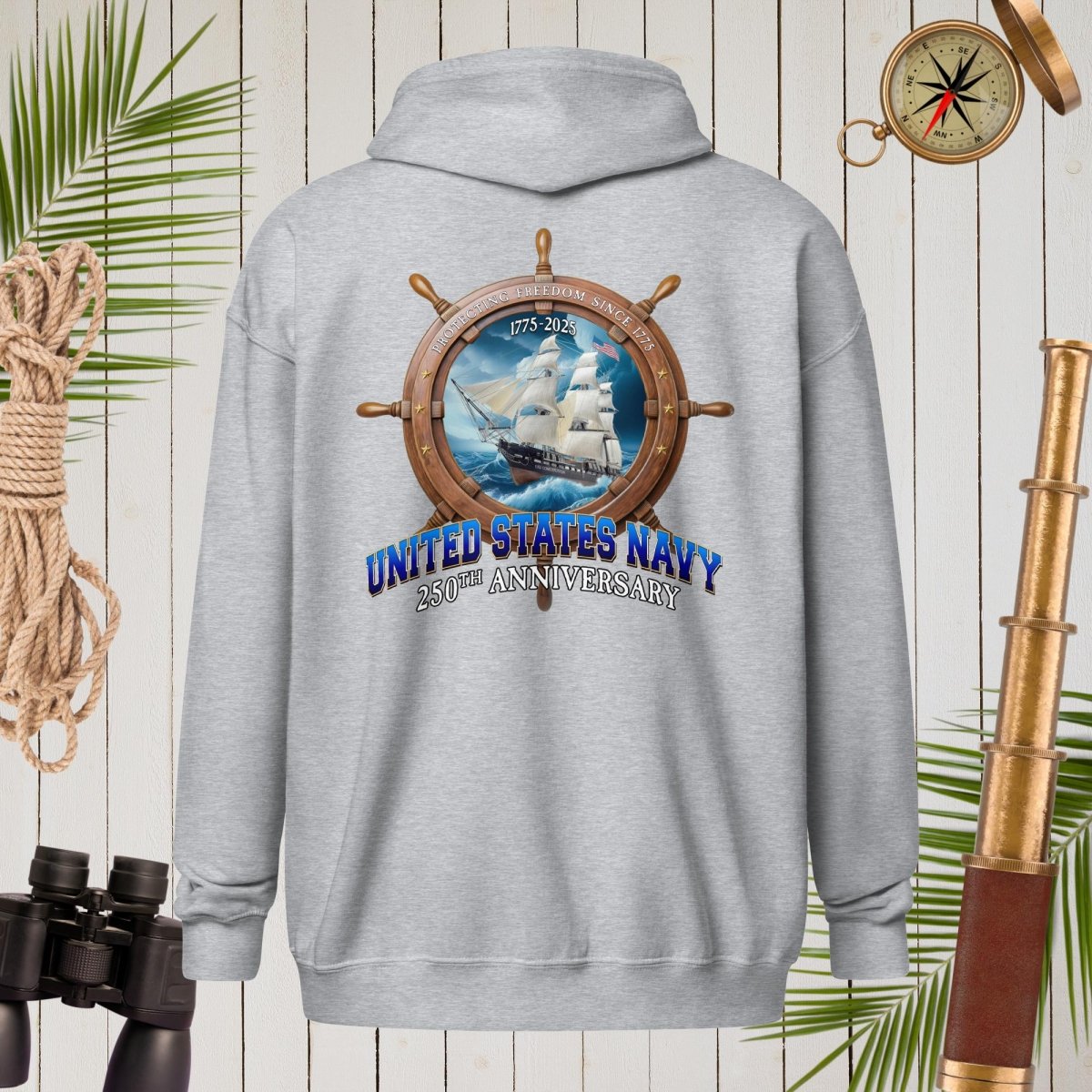 Gray hoodie with United States Navy 250th Anniversary design on a wooden background with nautical items.