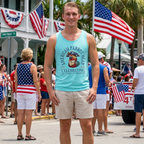 Man in a blue tank top with a patriotic ParrotHead design standing in a parade with American flags and people in the background.