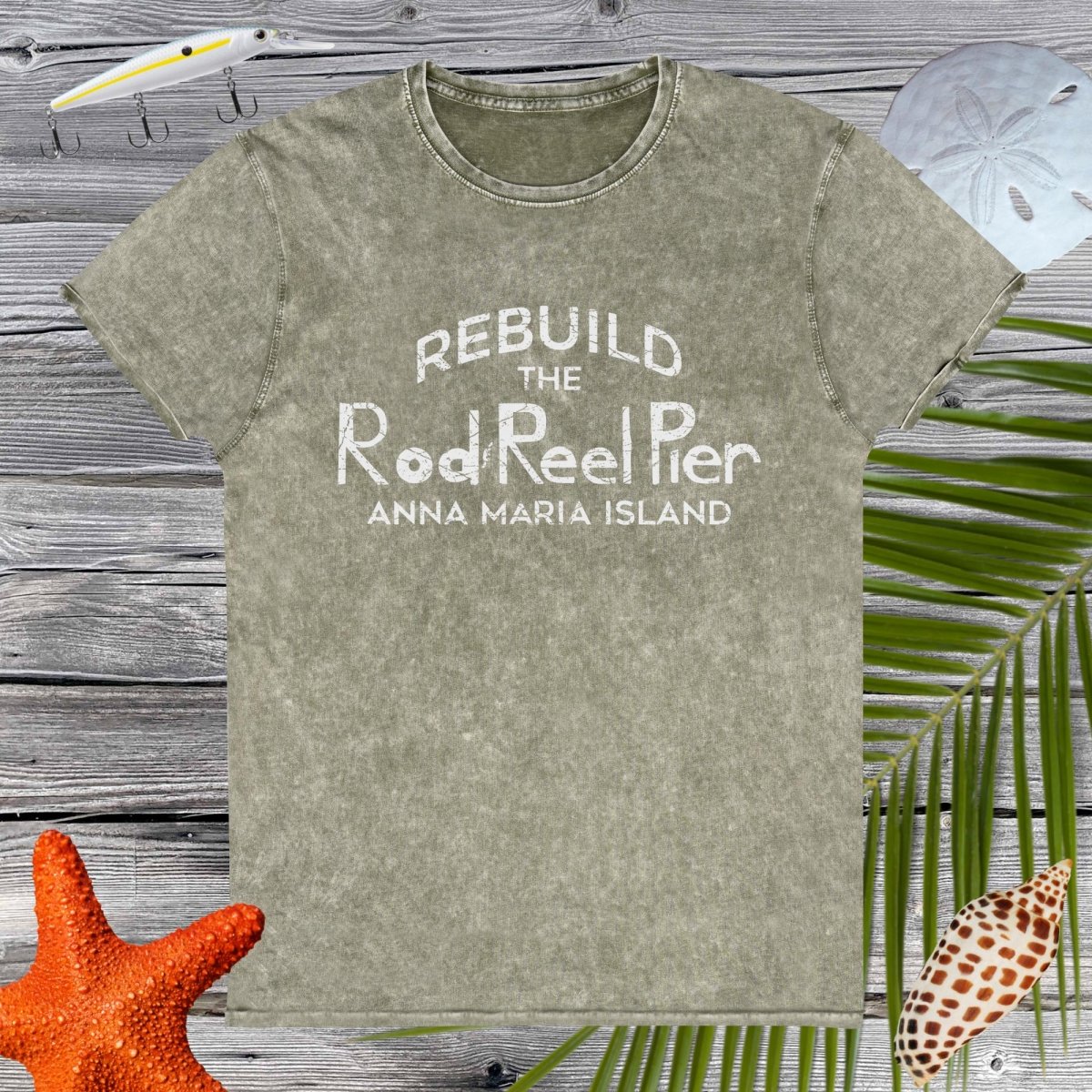 Green t-shirt with 'Rebuild the Rod & Reel Pier Anna Maria Island' text on a wooden surface with beach elements.