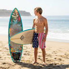 dude wearing American flag board shorts, holding a surfboard on La Jolla beach with a clear sky