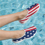 A pair of slip-on water shoes with an American flag pattern featuring white stars on a blue background and red and white stripes, shown on feet in a swimming pool.