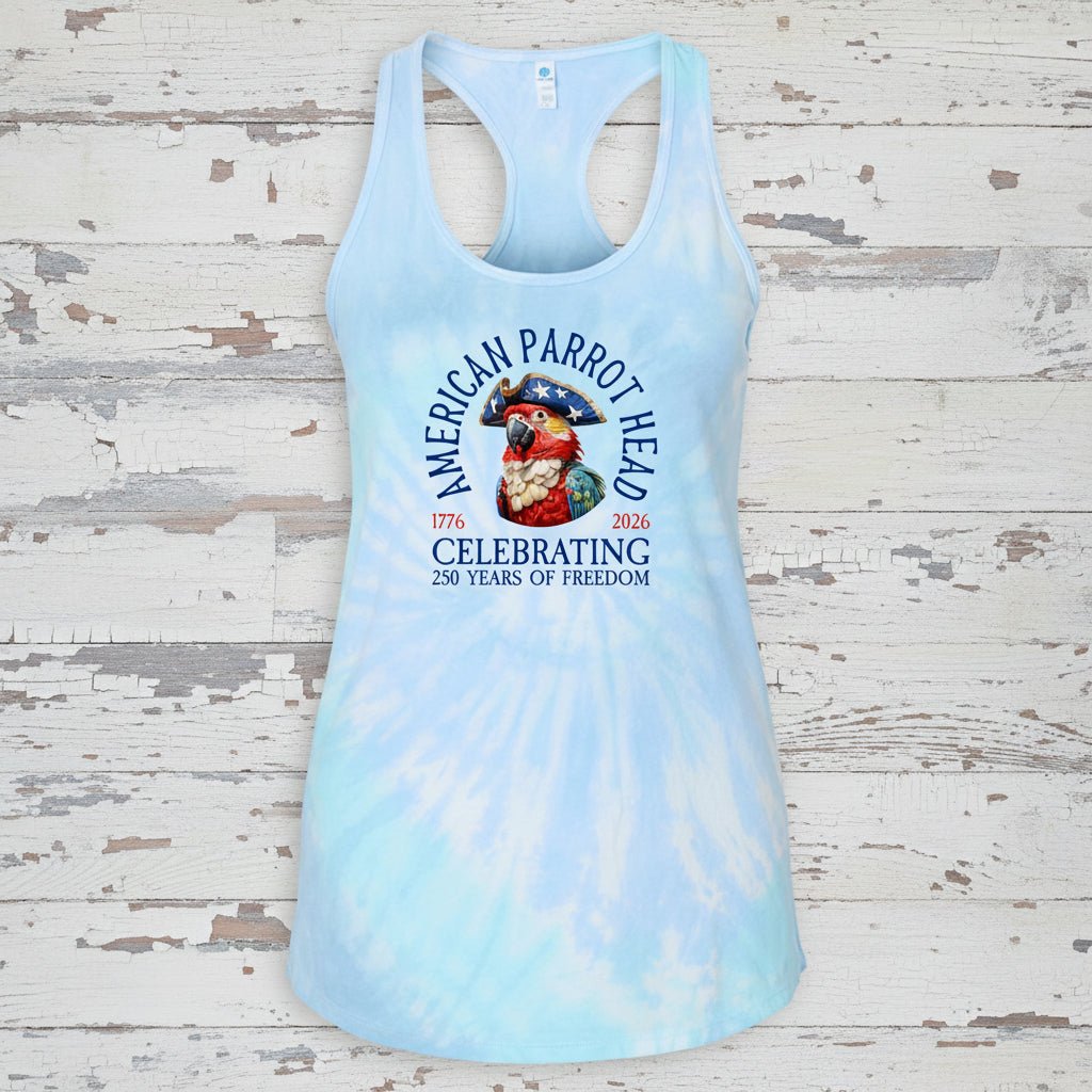 A light blue and white tie-dye racerback tank top featuring a 'American Parrot Head' 250th birthday graphic, displayed flat against a distressed white wood background.