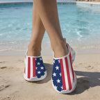 A person standing on a sandy beach near a swimming pool wearing Beach Walking Slippers with an American flag print featuring red and white stripes and white stars on a blue background.