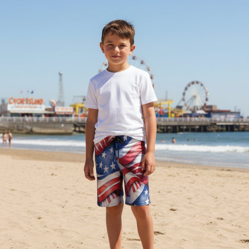 Boy on beach vacation wearing Kids Americana Board Shorts in front of an amusement park pier.