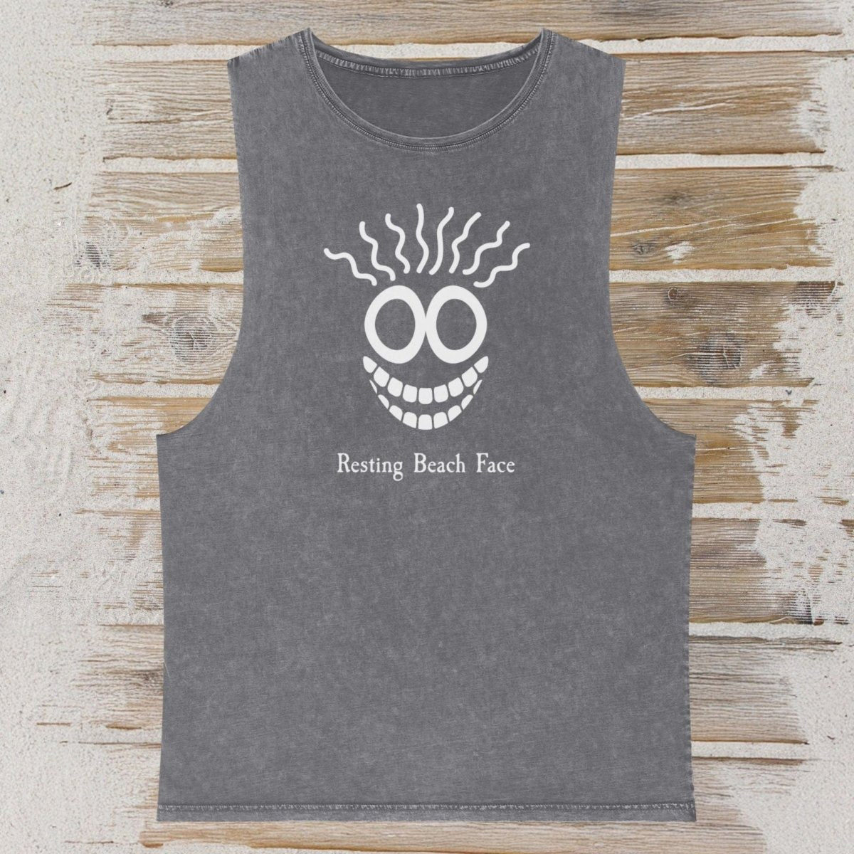 A dark gray sleeveless tank top with a white graphic of a skull-like face and the text 'Resting Beach Face' printed on the front, displayed flat against a rustic wood and beach sand