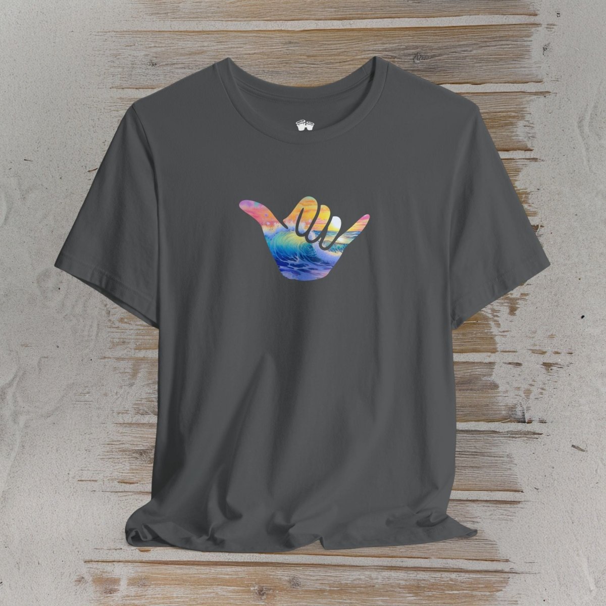A dark gray crewneck t-shirt featuring a colorful Hawaiian 'shaka' hand graphic on the chest, displayed flat against a rustic wooden plank background.