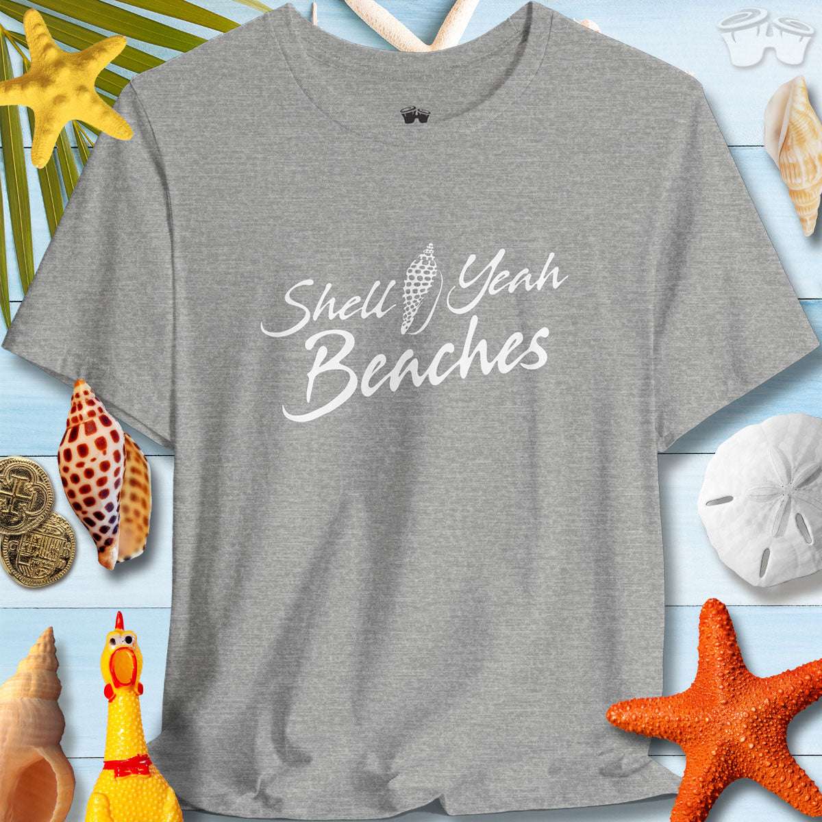 Gray short sleeve tee with 'Shell Yeah Beaches' text, surrounded by seashells and beach decor, promoting summer vibes.