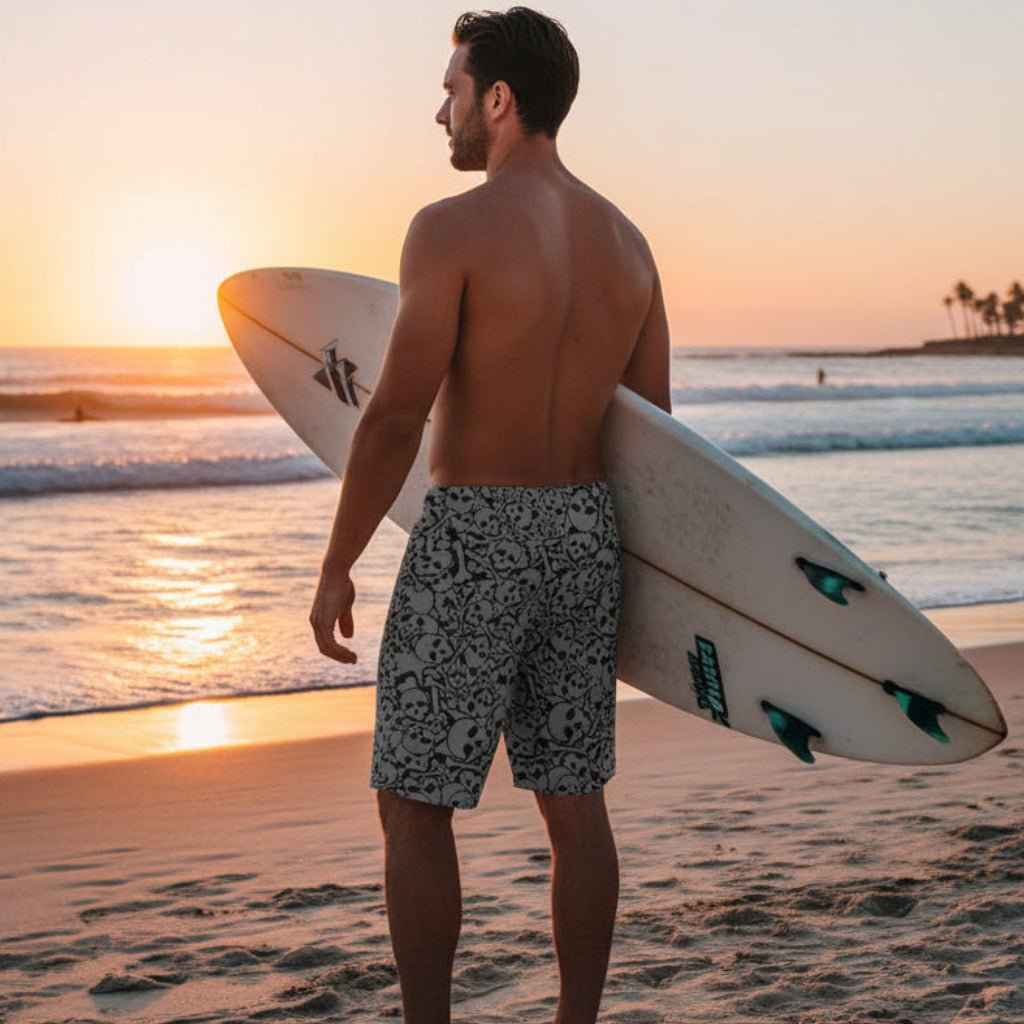 Guy wearing grey shorts with black skull pattern holding up a surfboard looking out at the ocean