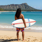 Pacific Islander dude with long hair wearing Colorful palm tree patterned board shorts standing on Waikiki Beach holding a surfboard and looking out at the waves