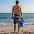 Dude wearing pirate pattern board shorts standing on a beach looking out at the ocean holding a pair of swim-fins, mask and snorkel 