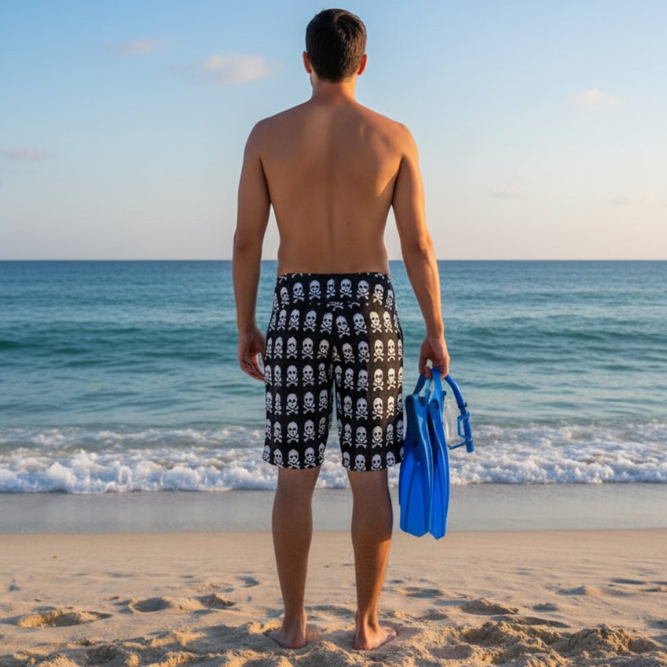 Dude wearing pirate pattern board shorts standing on a beach looking out at the ocean holding a pair of swim-fins, mask and snorkel 