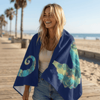 A woman smiling on a beach boardwalk wearing a navy blue wrap or towel with large tropical wave and palm tree prints in teal, aqua, and yellow-green. The beach, sand, and palm trees are visible in the background.