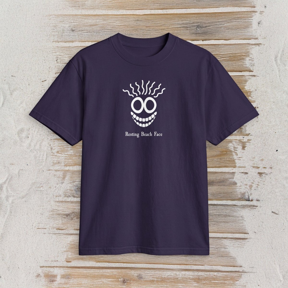 A dark purple short-sleeve t-shirt with a white graphic of a smiling face with wavy hair and the text 'Resting Beach Face' printed on the chest, displayed flat on a light wood plank background.