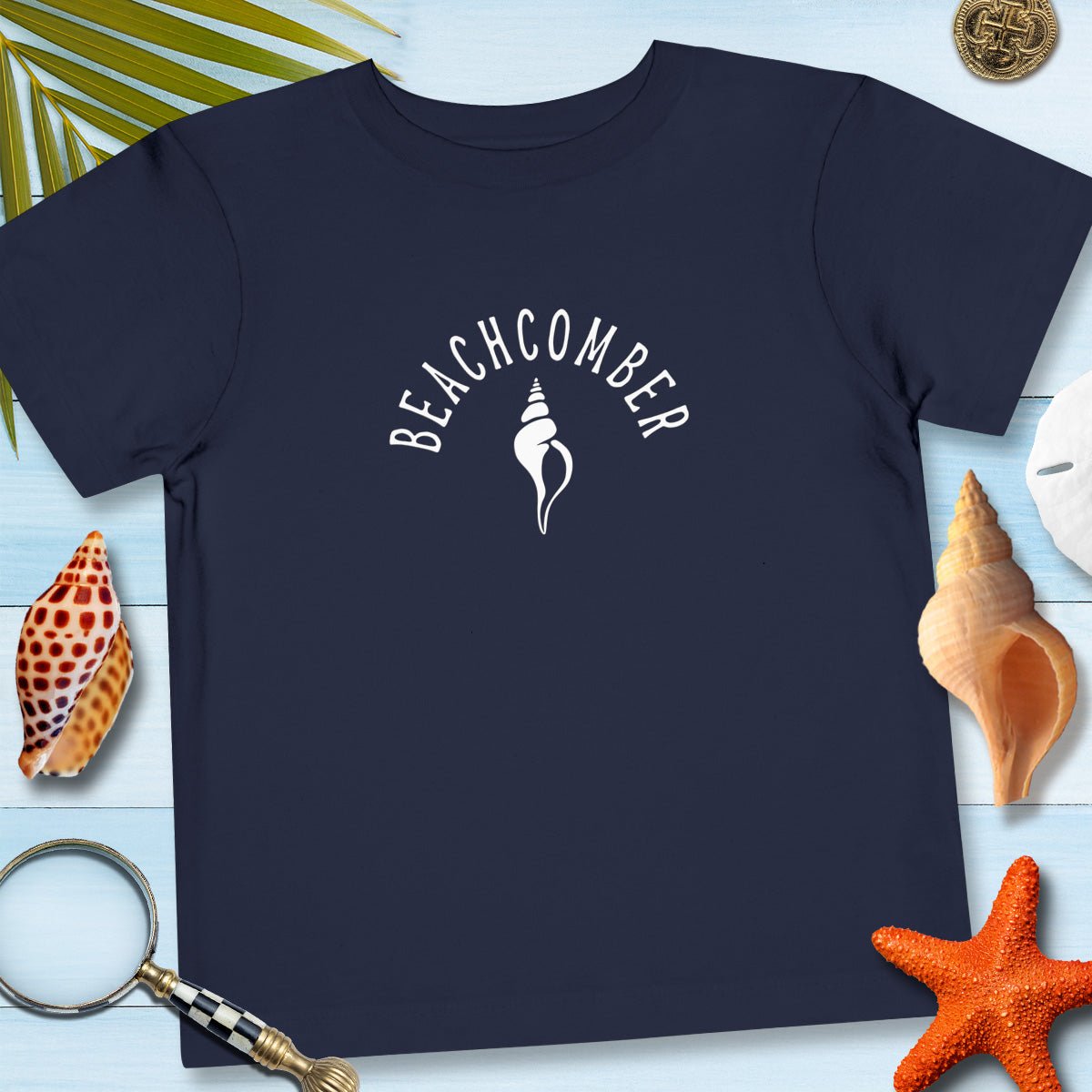 Navy blue t-shirt with 'BEACHCOMBER' design on a wooden surface with beach-themed items.