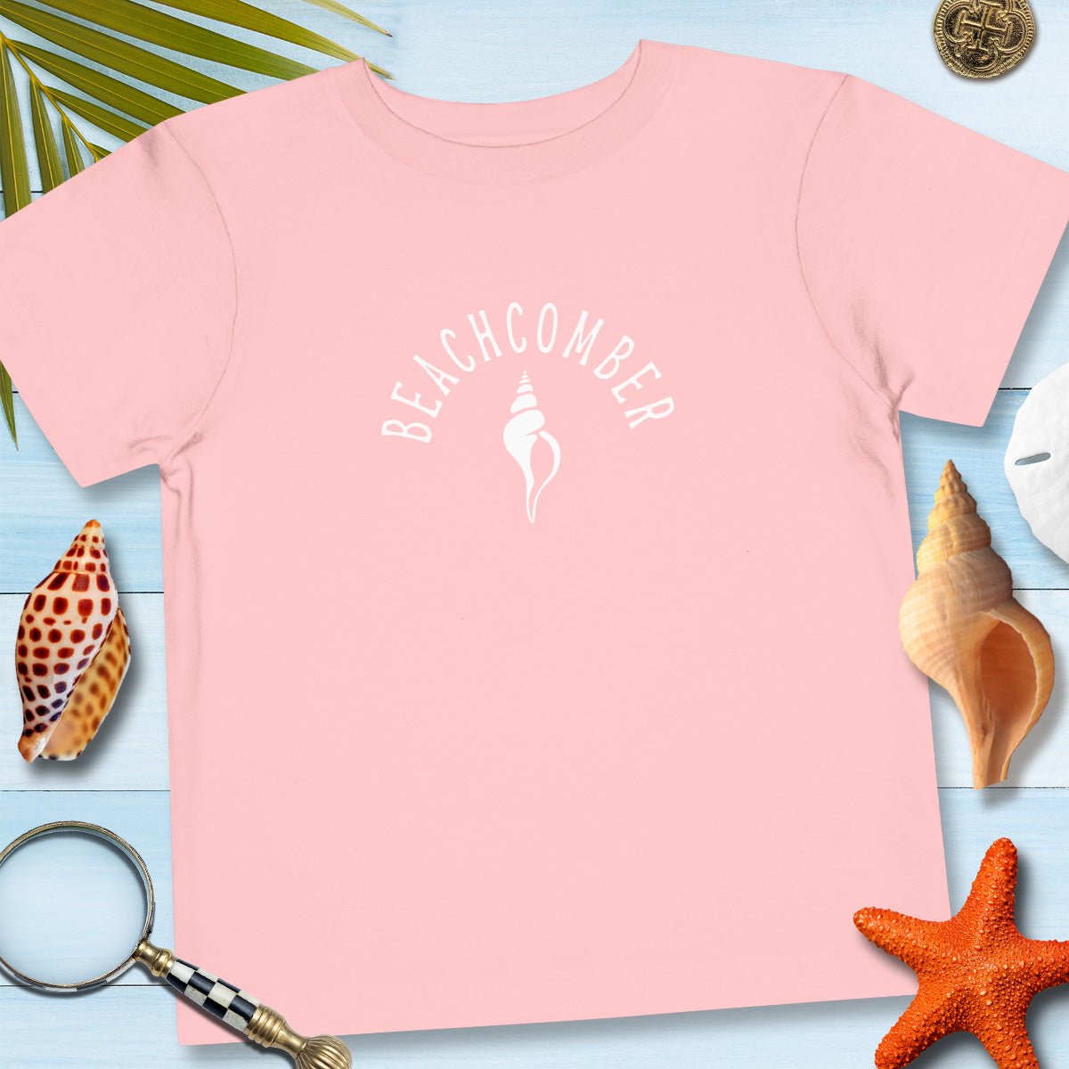 Pink t-shirt with 'BEACHCOMBER' text and shell graphic on a blue surface with beach-themed items.