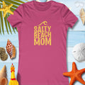 Women's Salty Beach Mom tee in pink with a gold print, surrounded by seashells and beach accessories.