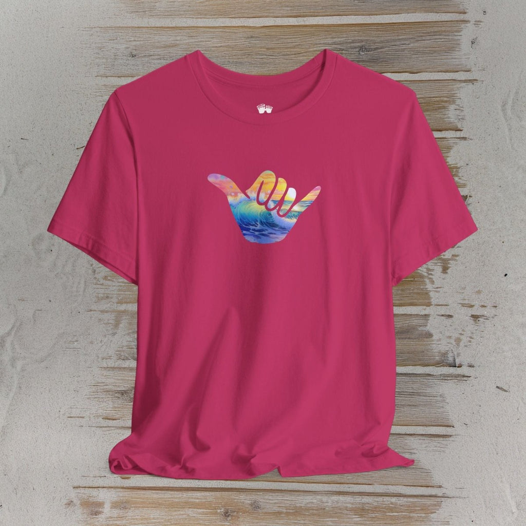 A bright pink short-sleeve t-shirt with a colorful graphic of a hand making the 'shaka' sign, filled with a sunset landscape. The t-shirt is displayed flat on a rustic, light-colored wooden plank background.