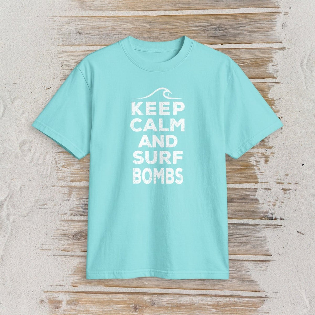 A light aqua blue short-sleeve t-shirt with a white graphic print reading 'KEEP CALM AND SURF BOMBS' on a rustic, light-colored wooden plank background.