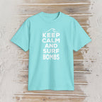 A light aqua blue short-sleeve t-shirt with a white graphic print reading 'KEEP CALM AND SURF BOMBS' on a rustic, light-colored wooden plank background.