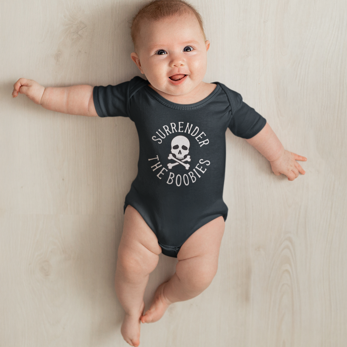 Baby wearing a dark gray onesie with text and a skull design on a light wooden floor.