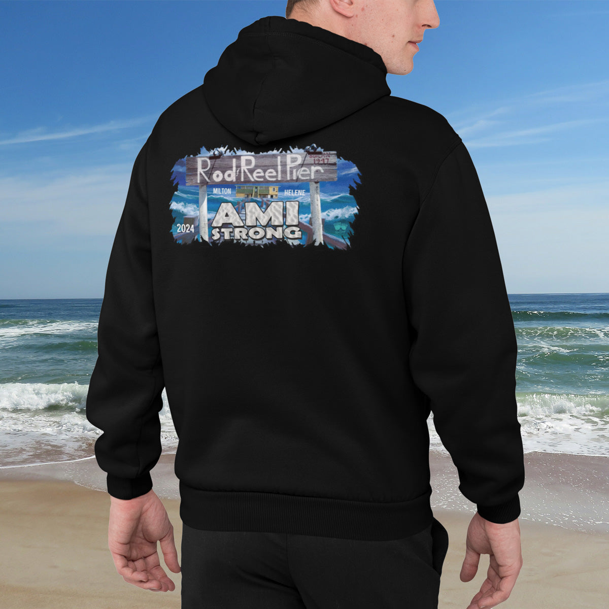 Person wearing a black hoodie with 'AMI Strong' design on a beach