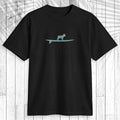 A black short-sleeve crewneck t-shirt featuring a light teal graphic of a dog standing on a surfboard on the chest, displayed flat against a white wood plank background with a leaf shadow overlay.