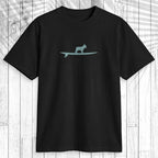 A black short-sleeve crewneck t-shirt featuring a light teal graphic of a dog standing on a surfboard on the chest, displayed flat against a white wood plank background with a leaf shadow overlay.