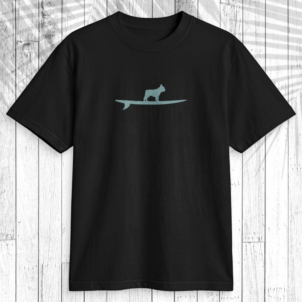 A black short-sleeve crewneck t-shirt featuring a light teal graphic of a dog standing on a surfboard on the chest, displayed flat against a white wood plank background with a leaf shadow overlay.