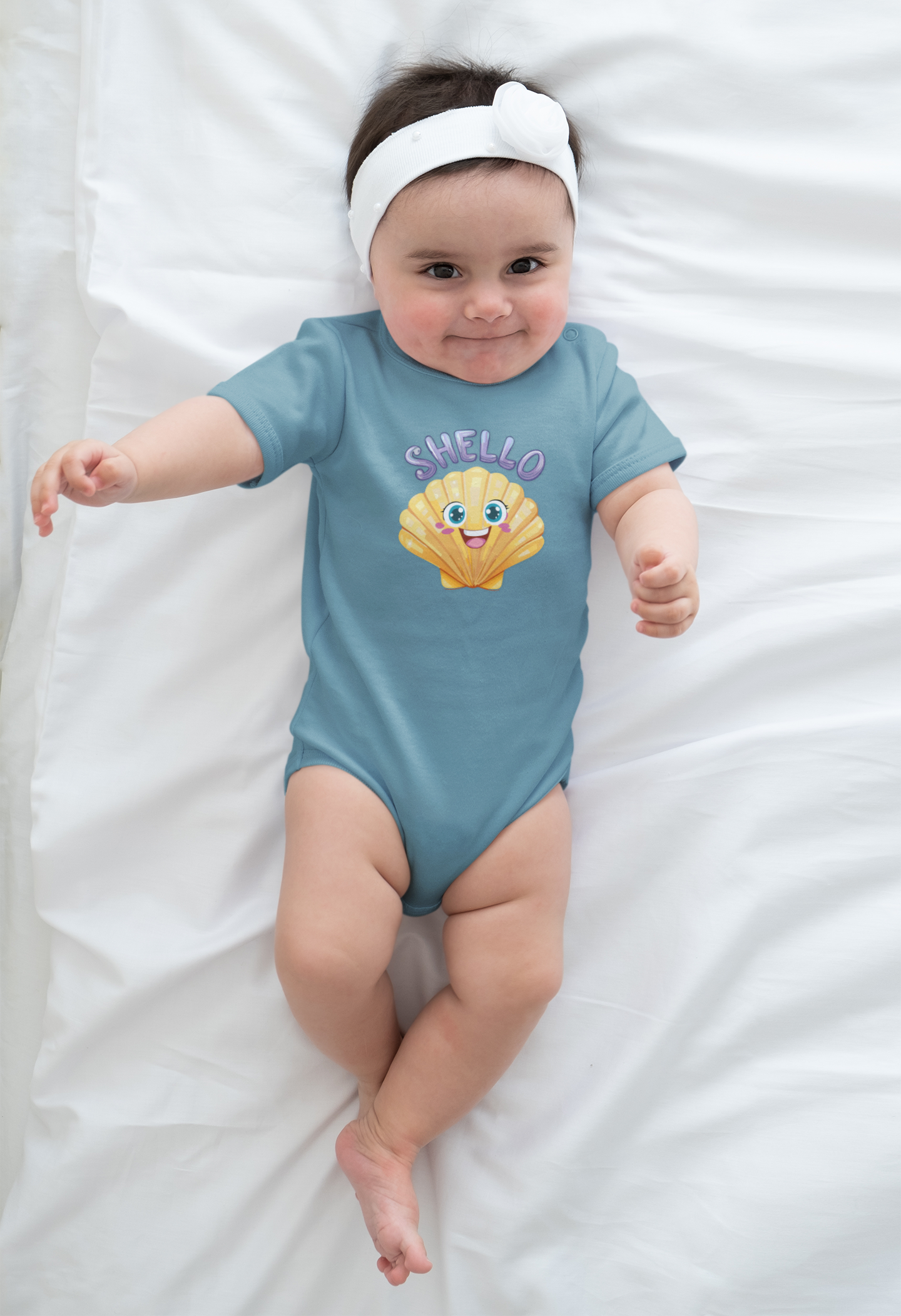 Baby wearing a blue onesie with a cartoon seashell character and Shello text, lying on a white blanket.