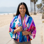 A woman standing on the boardwalk wearing a colorful Dolphin Print Beach Towel with blue, pink, purple, and white tones.