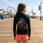 A girl viewed from behind wearing a black long-sleeve rash guard with a large colorful monster fish graphic on the back, paired with bright coral-orange shorts, standing on a sunny wooden boardwalk with a pier in the background.