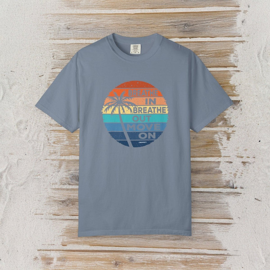 A slate blue crewneck t-shirt with a retro circular graphic on the chest, laid flat on a rustic white-washed wood background. The graphic features a sunset design with palm trees and the text 'MEAN IN BREATHE OUT MOVE ON'.
