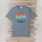 A slate blue crewneck t-shirt with a retro circular graphic on the chest, laid flat on a rustic white-washed wood background. The graphic features a sunset design with palm trees and the text 'MEAN IN BREATHE OUT MOVE ON'.