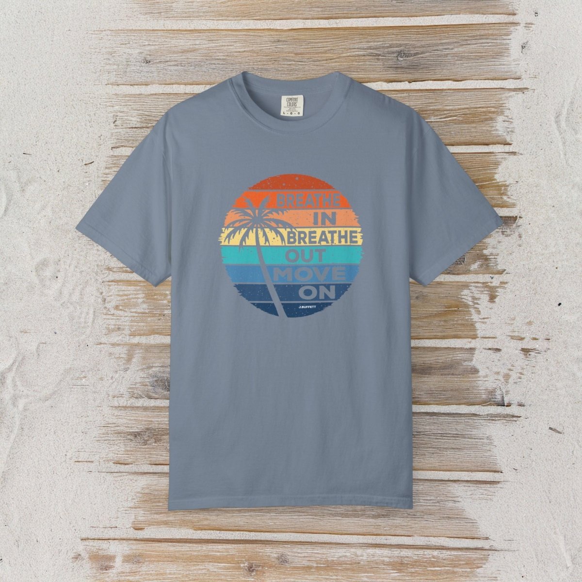 A slate blue crewneck t-shirt with a retro circular graphic on the chest, laid flat on a rustic white-washed wood background. The graphic features a sunset design with palm trees and the text 'MEAN IN BREATHE OUT MOVE ON'.