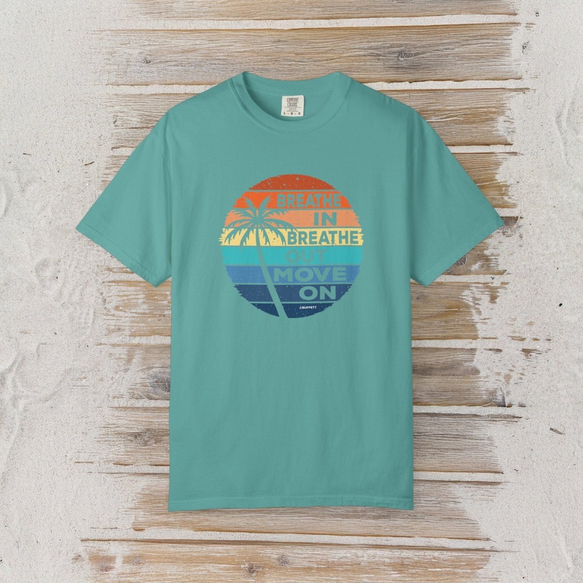A teal short-sleeve crewneck t-shirt with a retro circular graphic on the chest, laid flat on a rustic light wood plank background. The graphic features a sunset design with palm trees and the text 'Breathe IN BREATHE OUT MOVE ON'.