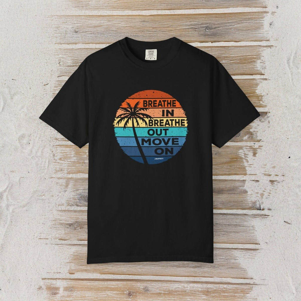 A black short-sleeve t-shirt with a retro circular graphic on the chest, laid flat on a light-colored wooden plank background. The graphic features a sunset design with palm trees and the text 'BREATHE IN BREATHE OUT MOVE ON' written in a vertical layout.