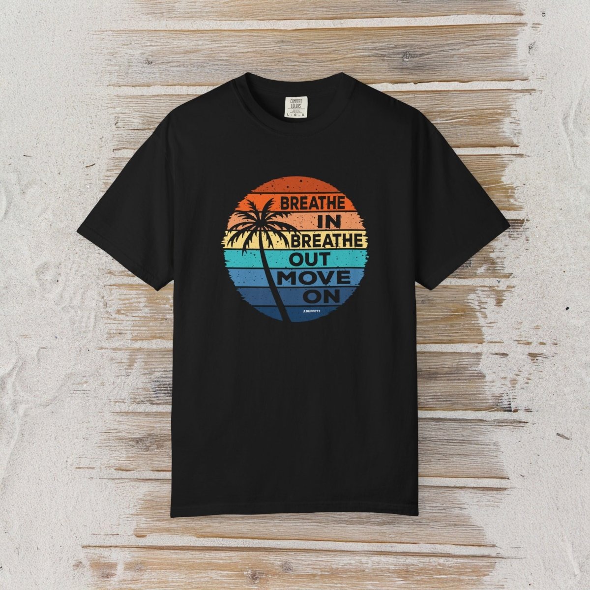 A black short-sleeve t-shirt with a retro circular graphic on the chest, laid flat on a light-colored wooden plank background. The graphic features a sunset design with palm trees and the text 'BREATHE IN BREATHE OUT MOVE ON' written in a vertical layout.