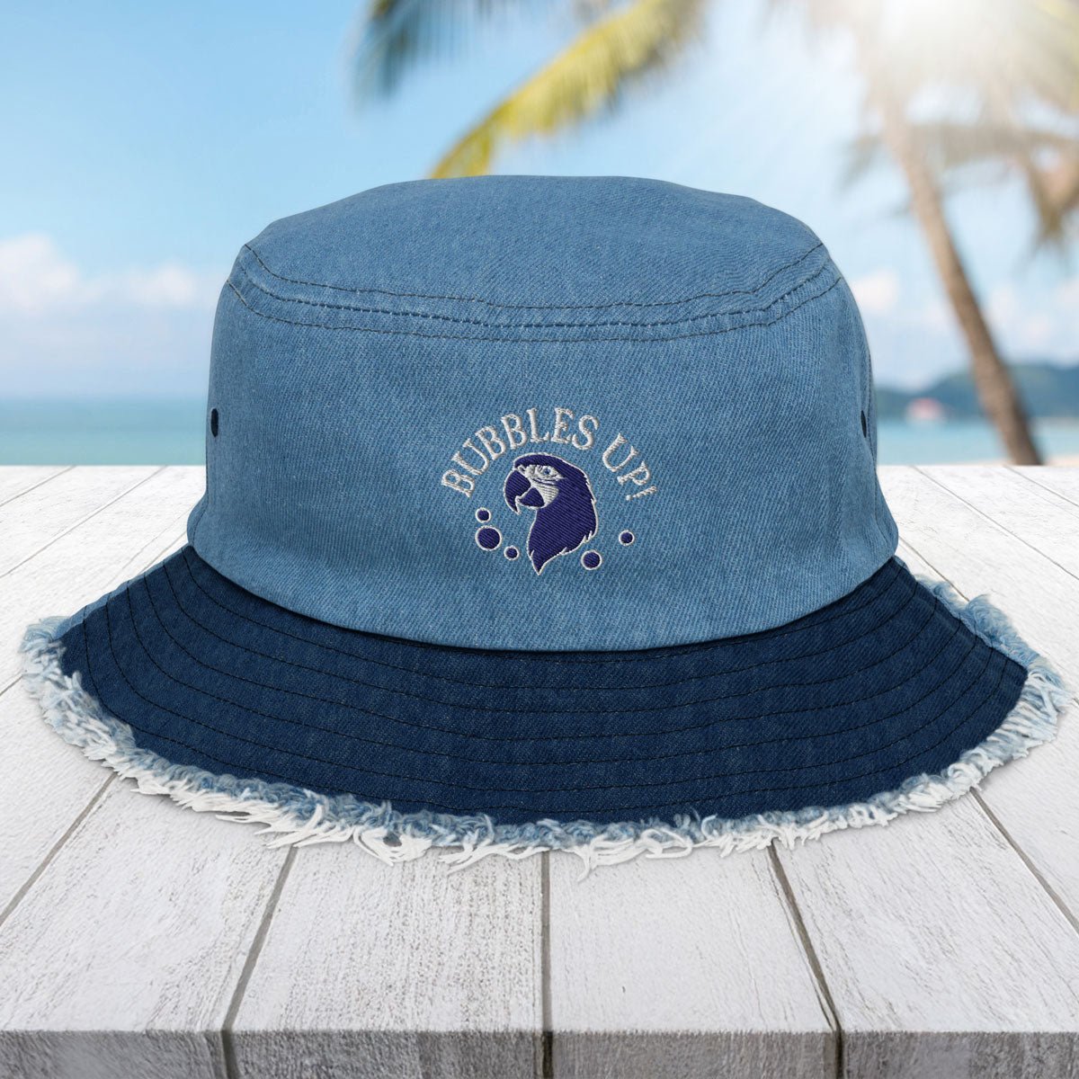 Blue bucket hat with 'Bubbles Up' logo on a wooden surface with a beach background