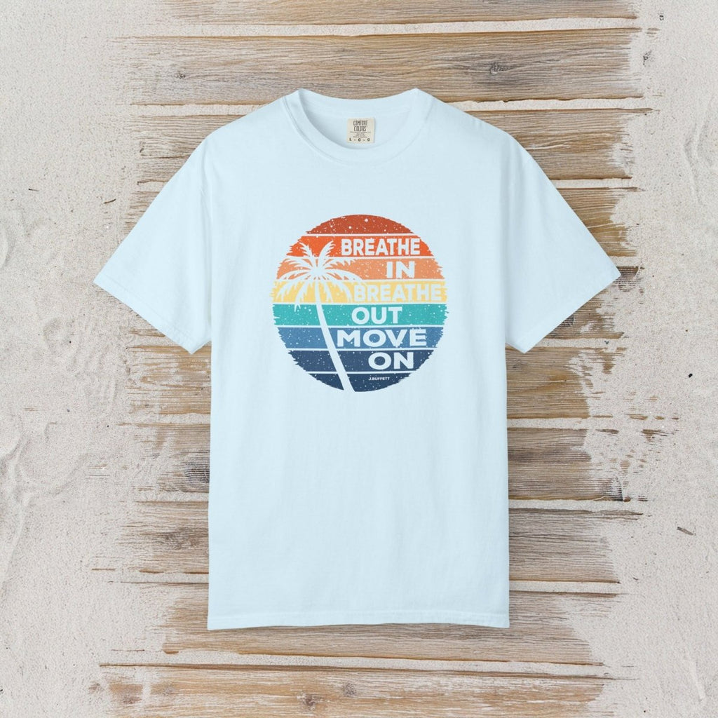 A light blue short-sleeve t-shirt with a circular retro graphic on the chest, laid flat on a rustic wooden plank background. The graphic features a sunset gradient in orange, yellow, green, and blue with a palm tree silhouette and the text 'BREATHE IN BREATHE OUT MOVE ON'.